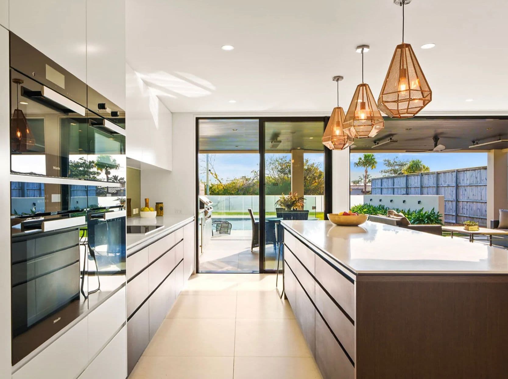 Handleless Sydney kitchen with a dark island, woven pendants and poolside sliding doors